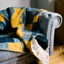 This quilt is stitched in a traditional strip design made from; sunshine yellow, grey and navy blue tartan Harris Tweed, trimmed with retro Hedgerow by renowned designer Angie Lewin and backed in a smoky grey velvet.
